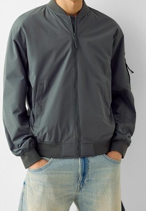 Gray bomber jacket made of smooth fabric, featuring a zip closure, ribbed cuffs, and side pockets. Worn over light blue jeans.