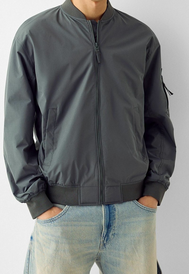 Grå bomberjacka i glatt tyg, med dragkedja, ribbade ärmslut och sidofickor. Bärs över ljusblå jeans.