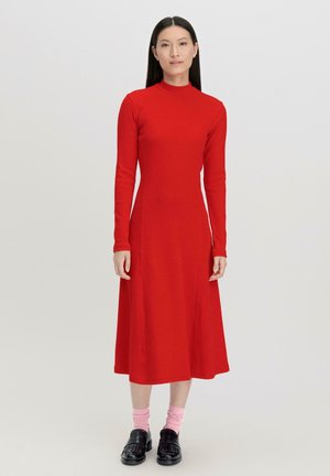Rotes, langärmliges Kleid mit hohem Ausschnitt, figurbetontem Oberteil und schwingendem, knielangem Rock, aus weichem Stoff. Kombiniert mit schwarzen Schuhen und rosa Socken.