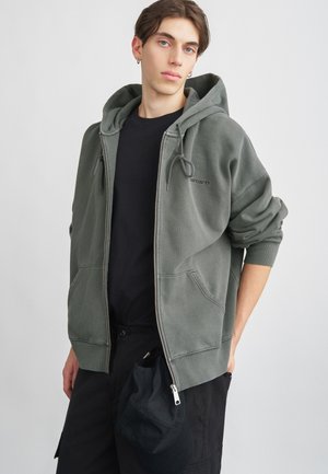 Olivgrüner Reißverschluss-Hoodie mit zwei vorderen Taschen, Kapuze mit Kordelzug und weicher Textur, getragen über einem schwarzen T-Shirt und kombiniert mit einer schwarzen Mütze.