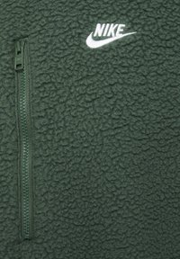 Casaco de fleece da Nike em verde escuro. Apresenta uma superfície texturizada, bolso com fecho zip no lado esquerdo do peito e logótipo bordado em branco.