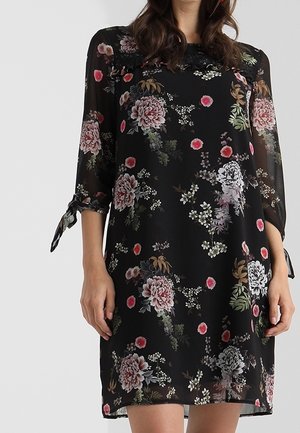 Zwarte jurk tot op de knie met een roze, witte en groene bloemenprint, doorschijnende driekwart mouwen die aan de manchetten zijn gebonden, gedragen door een staande persoon.