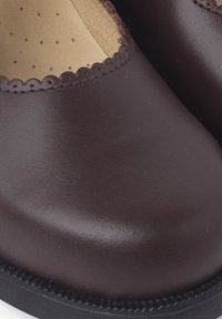 Zapato de cuero marrón con punta redondeada y borde ondulado. Presenta una textura suave y una suela de goma negra.