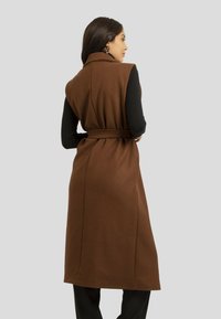 Manteau sans manches marron avec une taille ceinturée ; en tissu de laine texturé ; haut à manches longues noir contrastant en dessous ; coupe ajustée.