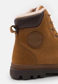 Botte en suède marron avec une doublure en peluche, un dessus en cuir marron renforcé et une semelle en caoutchouc texturé. Présente un logo circulaire et une tirette.