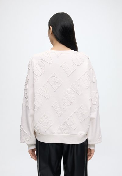Femme aux longs cheveux foncés porte un sweat-shirt blanc avec des lettres texturées "LOVE" sur tout le dos et un pantalon noir, de dos.