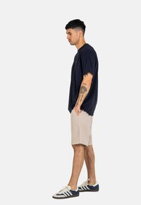Camiseta de algodón de color marino, corte relajado, combinada con pantalones cortos beige. El modelo lleva zapatillas deportivas blancas y marinas. Tatuajes visibles en los brazos.