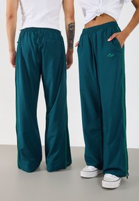 Teal Jogginghose mit lockerer Passform, grünen Seitenstreifen und elastischem Bund. Mit Taschen und Reißverschlüssen an den Säumen.
