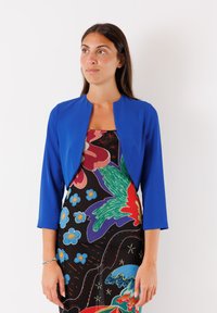 Giacca blu brillante cropped con maniche a tre quarti, indossata sopra un vestito colorato con motivo floreale su sfondo nero.