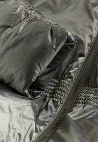 Tummanvihreä puhvitakki kiiltävästä, teksturoidusta kankaasta. Ominaisuuksiin kuuluu kerätty yksityiskohta ja logopatchi reunassa. Tiivis kolmiomainen muoto.