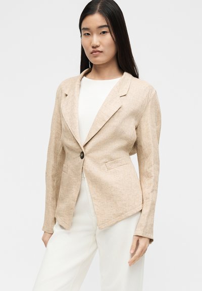 Γυναίκα με μπεζ σακάκι με ένα κουμπί πάνω από άσπρο τοπ και άσπρο παντελόνι, στέκεται μπροστά σε ένα απλό ανοιχτό φόντο.