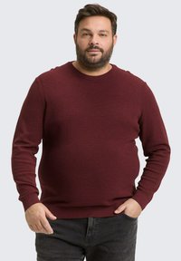 Burgunderfarbener Strickpullover mit Rundhalsausschnitt und langen Ärmeln. Elastisches Material mit texturierter Oberfläche. Kombiniert mit dunklen Jeans.