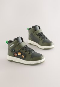 Sneakers alte in materiale sintetico verde oliva, con cinturino a strappo, dettagli neri e toppe decorative a tema videogioco.