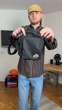 Μαύρη τσάντα χιαστί από ύφασμα με φερμουάρ, ρυθμιζόμενη λουρί και μικρή θήκη με λογότυπο στο μπροστινό μέρος. Συμπαγής ορθογώνια μορφή.