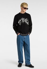 Suéter negro con diseño gráfico blanco, cuello y puños acanalados, combinado con jeans de denim azul holgados y zapatos negros. Lleva puestas gafas de sol.