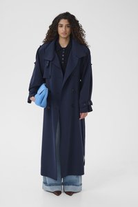 Frau mit langen lockigen Haaren, die einen marineblauen, doppelt geknöpften Trenchcoat, weit umgeschlagene Jeans, braune spitze Schuhe trägt und eine blaue Clutch hält.