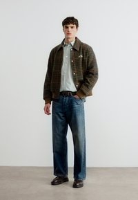 Νεαρός άνδρας με καφέ καρό σακάκι, ανοιχτό γκρι πουκάμισο, μπλε τζιν και σκούρα καφέ παπούτσια, στέκεται μπροστά σε λευκό φόντο.