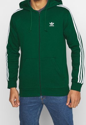 Muž v tmavě zelené mikině značky Adidas na zip s bílými pruhy na rukávech a modrých džínách, ruce volně podél těla.