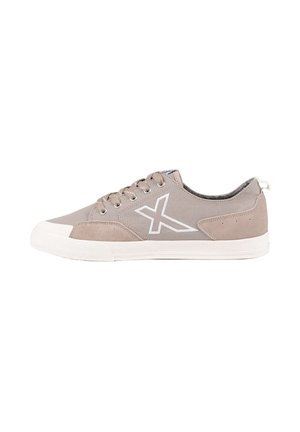 Zapatilla baja de lona color topo con suela blanca, cordones frontales, detalles de ante en la puntera y el talón, y un logo X blanco cosido en el lateral.