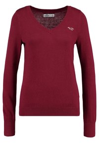 Burgundy stickad tröja med långa ärmar, V-ringning, ribbade ärmslut och nederkant. Litet, vitt logotypdetalj på vänster bröstområde. Storlek S.