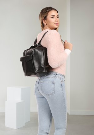 Frau trägt hellrosa flauschigen Pullover und hellblaue Jeans, trägt schwarzen Leder-Rucksack über der Schulter in einem schlichten Raum.