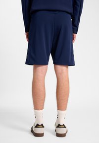 Marineblaue Sportshorts mit glatter Textur, ausgestattet mit einem taillierten Bund und reflektierenden Akzenten an der Seite. Kombiniert mit Sneakers und Socken.