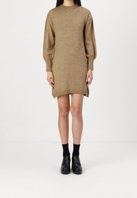 Robe en maille couleur fauve avec une coupe droite, des poignets et un ourlet légèrement côtelés. Dotée d'un col montant et de manches ballon. Associée à des chaussures et des chaussettes noires.