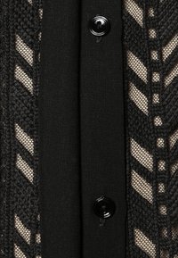 Indumento nero con bottoni, caratterizzato da un motivo a maglia testurizzata e a rete. Il design include una sezione di tessuto nero uniforme adiacente alla maglia.