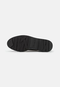 Semelle de chaussure en caoutchouc noir avec des motifs de grip texturés, dotée d'un logo circulaire et de rainures diagonales pour l'adhérence et la stabilité.