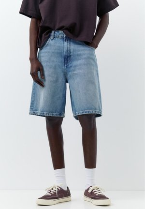 Osoba nosząca jasnoniebieskie szorty jeansowe, ciemną koszulę częściowo wpuszczoną w spodnie, białe skarpetki sportowe oraz ciemnoczerwone tenisówki z białymi sznurówkami.