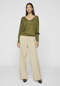 Olijfgroene V-hals trui met een losse pasvorm en lange mouwen, gecombineerd met beige wijde broekspijpen. De outfit bevat subtiele textuurdetails.