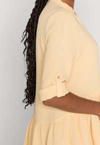 Light yellow, textured dress with buttoned cuff sleeves and a tiered skirt design, featuring a collared neckline and gathered fabric details.