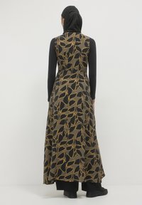 Langes Kleid in schwarz mit goldenen Blumenmuster, aus leichtem Stoff gefertigt. Es hat ein enganliegendes Oberteil und einen fließenden Rock, kombiniert mit langen schwarzen Ärmeln.