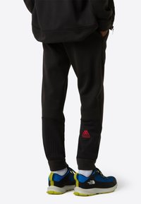 Svarte treningsbukser med ribt tekstur nederst på benet, med en rød logo. Kombinert med blå og svarte joggesko med knallgule aksenter.