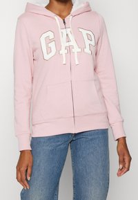 Donna che indossa una felpa con zip color rosa chiaro con le lettere bianche GAP sul petto e jeans blu, in piedi contro uno sfondo neutro.