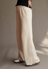 Beige afslappede bukser med vidde, elastisk talje og trykknapsåbninger i siden. Glat tekstur, parret med hvide sneakers.
