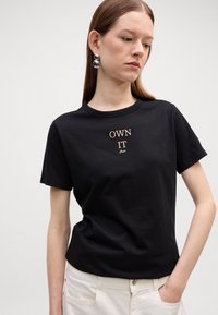Svart bomullst-skjorte med kort erme, med brodert tekst "OWN IT" i gull. Modellen har på seg hvite bukser, som viser en enkel design.