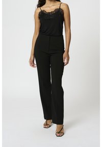 Camisola nera con applicazioni in pizzo abbinata a pantaloni neri sartoriali, con vita alta e gamba dritta, completati da sandali neri con cinturini.