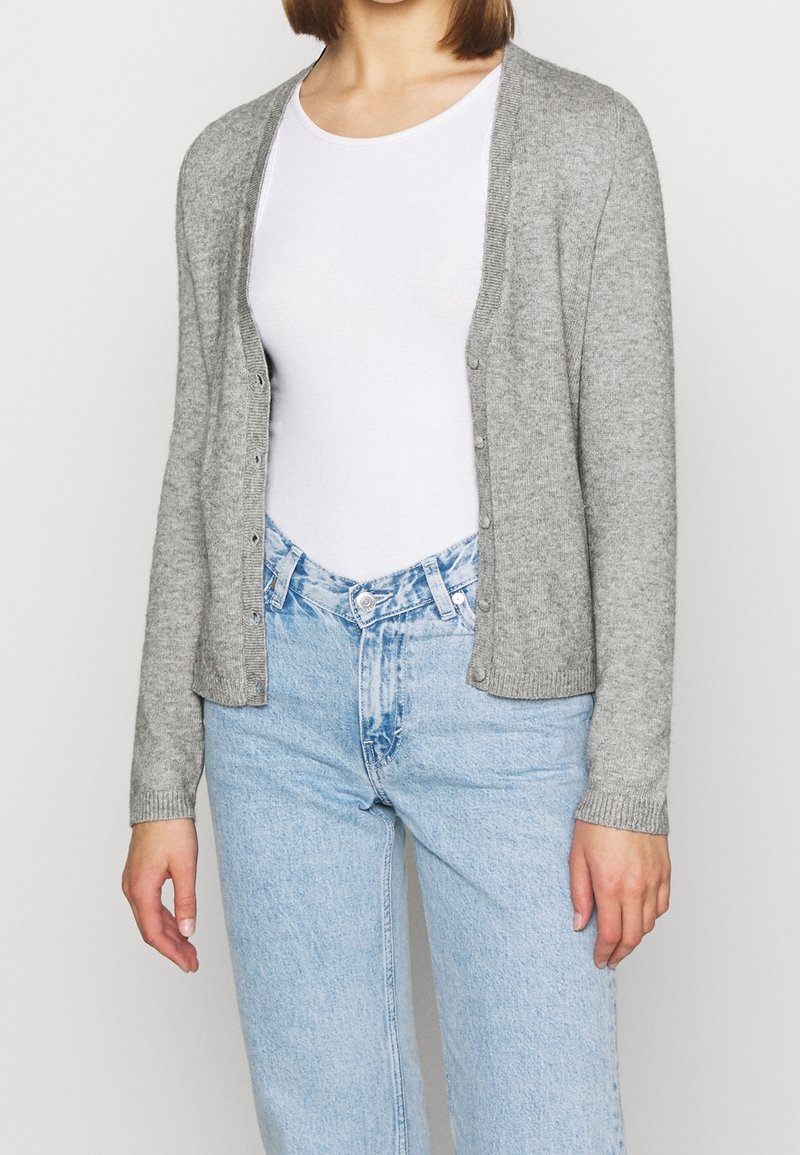 Personne portant un cardigan gris clair boutonné sur un t-shirt blanc associé à un jean taille haute bleu clair, debout avec les bras détendus.