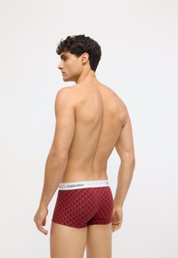 Czerwone bokserki o geometrycznym wzorze, wykonane z miękkiego materiału, z białym logo Calvin Klein na pasku. Widok z tyłu.