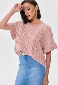 Różowy sweter z dzianiny o splocie w warkocze, krótkie rękawy i luźny krój, połączony z jasnoniebieską spódnicą dżinsową.