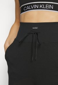 Zbliżenie na kobietę w czarnym biustonoszu sportowym Calvin Klein oraz czarnych spodniach z wysokim stanem, z jawą i przednim logo Calvin Klein.