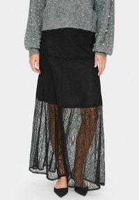 Gonna maxi in pizzo nero con sovrapposizione trasparente. Presenta un orlo scallop e un design a strati. Indossata con un maglione grigio e ballerine a punta.