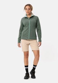 Giacca verde con zip e un motivo testurizzato, abbinata a pantaloni corti beige. Calze nere con strisce bianche e scarpe da escursionismo nere.
