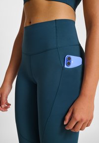 Tealfärgade högmidjade leggings i stretchigt tyg, med en sidoficka som håller en ljusblå smartphone. Slät yta och figurnära design.