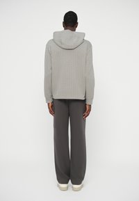 Άνδρας που στέκεται με την πλάτη γυρισμένη, φορώντας γκρι πλεκτή κουκούλα, σκούρους γκρίζους φαρδιούς παντελόνες με τσέπη στο πλάι, και μπεζ αθλητικά παπούτσια σε λευκό φόντο.