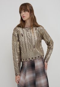 Pull en or métallique avec un design en tricot à torsades, encolure ronde et ourlet côtelé. Porté par-dessus une jupe à carreaux dans des tons de marron et de noir atténués.