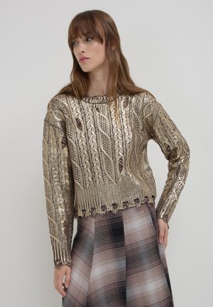 Metallisch goldener Pullover mit Zopfmuster, rundem Halsausschnitt und geripptem Saum. Über einem karierten Rock in gedämpften Brauntönen und Schwarz getragen.