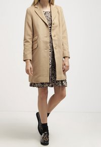Beżowy wełniany płaszcz z klapami, trzema guzikami i bocznymi kieszeniami, noszony na sukience w panterkę i czarnych butach.