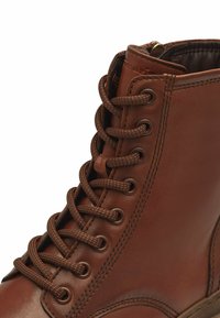 Bottines en cuir marron avec une surface texturée, bout arrondi et lacets assortis. Elles présentent une fermeture éclair sur le côté et des détails de couture contrastants.